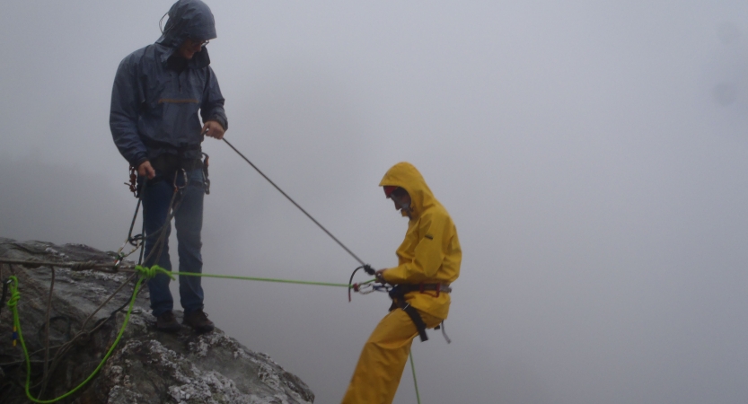 Two people wearing safety gear are secured by ropes near the edge of a cliff. One person appears to be an instructor, giving direction to the other person.